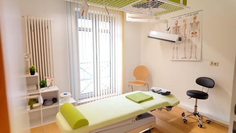 Modern physical therapy clinic room with treatment table, exercise equipment, anatomical wall chart, and bright natural lighting from large windows