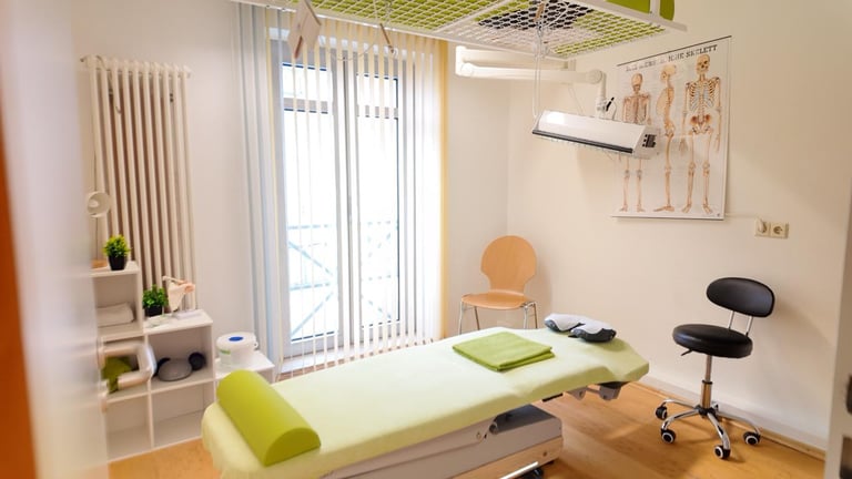 Bright physical therapy clinic room with treatment table, exercise equipment, anatomical charts, and natural lighting from large windows